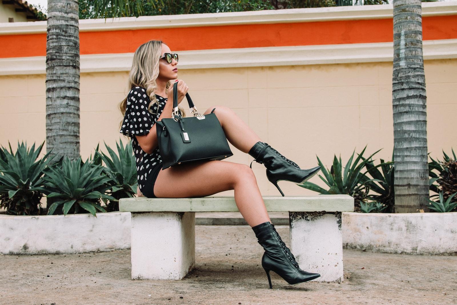 Woman Wearing Black and White Polka-dot Shirt With Black Short Shorts Holding Black Leather Tote Bag Sitting on White Concrete Bench